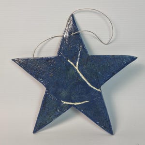 Op de afbeelding: Een blauw keramisch stervormig ornament met een getextureerde afwerking en een ophanglus. De ster heeft witte craquelé details over het oppervlak. Dit decoratieve stuk is ideaal voor feestelijke of dagelijkse woondecoratie. Ongeveer 15 cm breed.