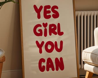 May include: A framed print with the words "YES GIRL YOU CAN" in red, bold letters. The print is displayed in a living room setting, next to a potted plant and a beige sofa. A window with a view of a building is in the background.