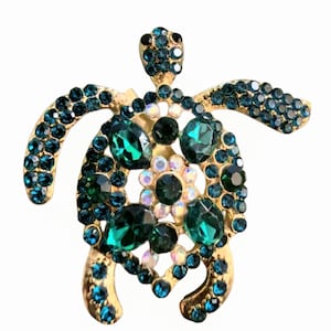 May include: A gold-toned turtle brooch with a detailed design. The brooch features a body of large, emerald green and smaller teal-colored rhinestones. The turtle's shell is adorned with a cluster of iridescent stones.