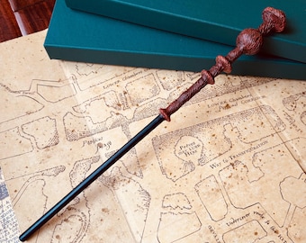 Op de afbeelding: Een zwarte en bruine toverstaf met een gedetailleerd handvat ligt op een kaart van Zweinstein. De kaart is bedrukt met de namen van verschillende locaties, waaronder "Lower Chambers", "Porticus Mage" en "Way to Transfiguration".