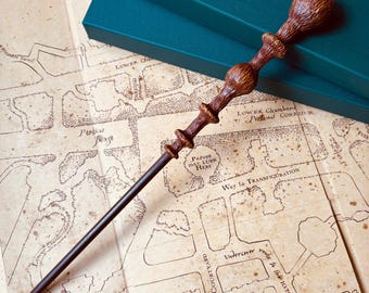May include: A brown wooden wand with a black metal core, lying on a map of Hogwarts. The map shows the location of "Peeves may Lurk Here" and "Way to Transfiguration".