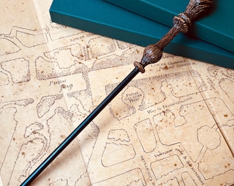 Op de afbeelding: Een zwarte en bruine toverstaf met een houten handvat en een zwarte schacht. De toverstaf ligt op een kaart met tekst die "Lower Chambers", "Peéves may LURK HERE" en "Way to TRANSFIGURATION" bevat.