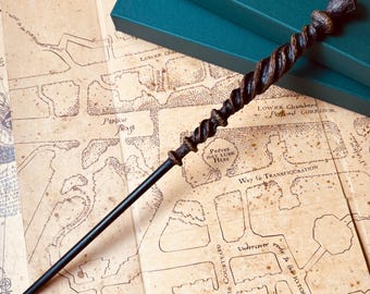 Op de afbeelding: Een donkerbruine en zwarte toverstok met een gedraaid ontwerp, liggend op een kaart van Zweinstein. De kaart toont de indeling van het kasteel, inclusief de locatie van de Kamer van Hoge Nood, de Zuiveringsleer-klas en de Grote Zaal.