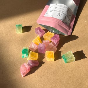 Può includere: Un sacchetto rosa aperto, etichettato "CRYSTALICIOUS SNACKS FRUIT MIX", con caramelle colorate a forma di cristallo che fuoriescono. Le caramelle sono di vari colori, tra cui rosa, giallo, viola e verde. Lo sfondo è una superficie marrone chiaro.