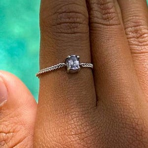 Puede incluir: Un anillo de plata con una piedra preciosa cuadrada y transparente. El anillo tiene una banda delicada y retorcida. El anillo se muestra sobre un fondo turquesa borroso.