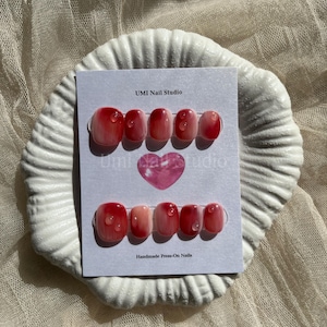 Puede incluir: Dos juegos de uñas postizas con un diseño degradado rojo y rosa, cada uno con detalles de gotas de agua. La tarjeta dice "UMI Nail Studio" y "Handmade Press-On Nails" con un corazón rosa.