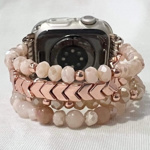 May include: A silver Apple Watch with three stacked bracelets in pink and rose gold tones. The bracelets include faceted and round beads, plus a chevron pattern. The watch and bracelets are presented on a white surface.