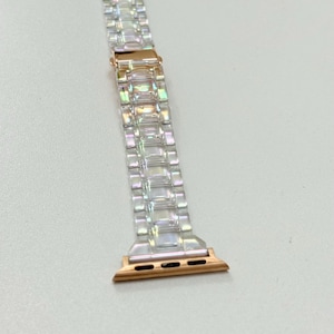 May include: A clear, iridescent watch band with a rose gold clasp and connectors. The band is made of rectangular links that catch the light, creating a shimmering effect. The connectors have three small holes for attachment.