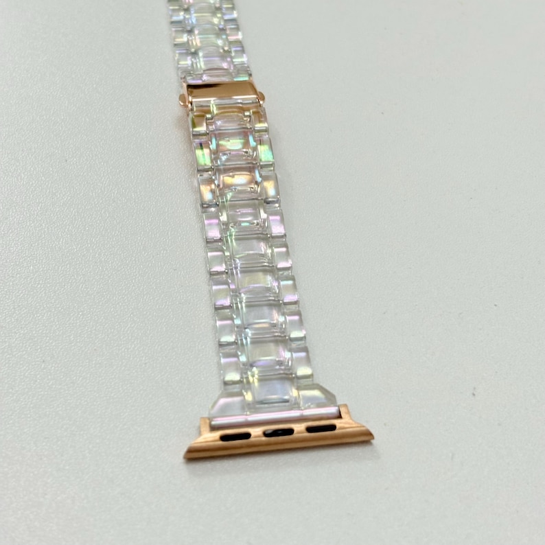 May include: A clear, iridescent watch band with a rose gold clasp and connectors. The band is made of rectangular links that catch the light, creating a shimmering effect. The connectors have three small holes for attachment.