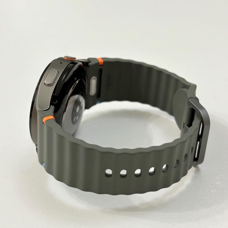 May include: A smartwatch with a dark grey case and a dark green, textured strap. The watch has orange detailing and a black screen. The strap is secured with a dark grey buckle.