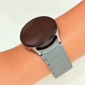 May include: A silver smartwatch with a gray band is worn on a wrist. The watch face is dark brown, and the band has a rectangular link design. The watch has an orange accent on the side.