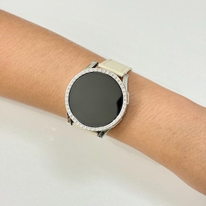 May include: A silver smartwatch with a black screen and a bezel encrusted with small, clear crystals. The watch has a cream-colored leather band. The watch is on a wrist.