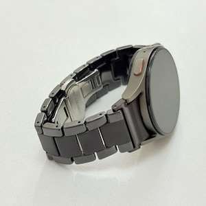 K&ouml;nnte beinhalten: Eine dunkelgraue Smartwatch mit passendem Gliederarmband. Das Zifferblatt der Uhr ist rund und dunkel, mit einer silberfarbenen L&uuml;nette. Das Armband besteht aus rechteckigen Gliedern. Die Uhr liegt auf einer wei&szlig;en Oberfl&auml;che.