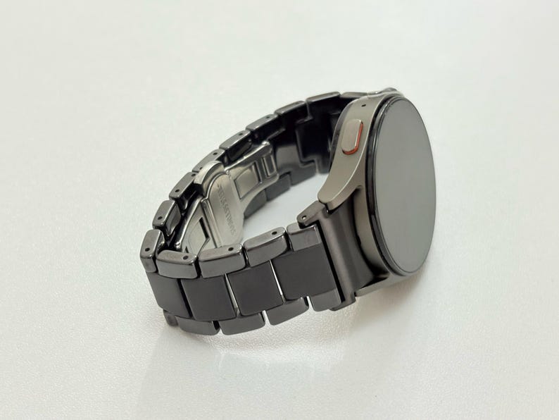 K&ouml;nnte beinhalten: Eine dunkelgraue Smartwatch mit passendem Gliederarmband. Das Zifferblatt der Uhr ist rund und dunkel, mit einer silberfarbenen L&uuml;nette. Das Armband besteht aus rechteckigen Gliedern. Die Uhr liegt auf einer wei&szlig;en Oberfl&auml;che.