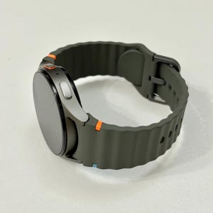 May include: A smart watch with a dark grey face and a dark olive green strap. The strap has a textured, ridged design and a silver buckle. Orange accents are visible on the watch casing. The watch is designed for fitness and activity tracking.
