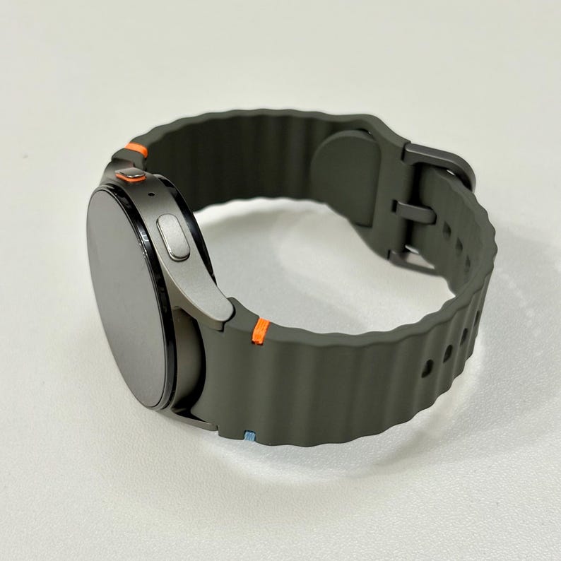 May include: A smart watch with a dark grey face and a dark olive green strap. The strap has a textured, ridged design and a silver buckle. Orange accents are visible on the watch casing. The watch is designed for fitness and activity tracking.