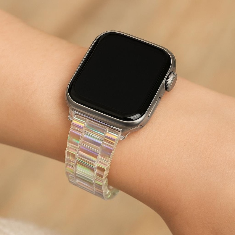 May include: A silver smartwatch with a clear, iridescent band is displayed on a wrist. The band is made of rectangular links that catch the light, creating a rainbow effect. The watch face is black.
