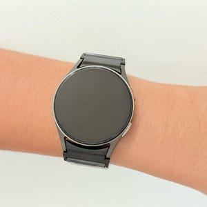 K&ouml;nnte beinhalten: Eine runde, schwarze Smartwatch mit einem dunkelgrauen Metallarmband wird am Handgelenk einer Person getragen. Das Zifferblatt der Uhr ist ein dunkler Bildschirm. Das Armband besteht aus rechteckigen Gliedern. Die Uhr ist ein modernes Accessoire.