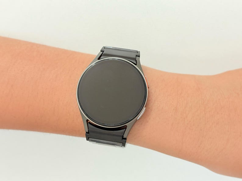 K&ouml;nnte beinhalten: Eine runde, schwarze Smartwatch mit einem dunkelgrauen Metallarmband wird am Handgelenk einer Person getragen. Das Zifferblatt der Uhr ist ein dunkler Bildschirm. Das Armband besteht aus rechteckigen Gliedern. Die Uhr ist ein modernes Accessoire.