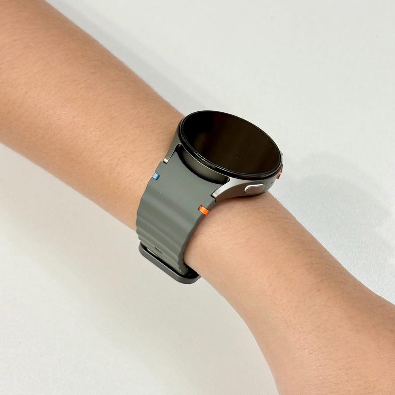 May include: A dark grey smartwatch with a black screen is worn on a wrist. The watch has a ribbed, dark grey band with small blue and orange accents. The watch face is round and the watch is on a light background.