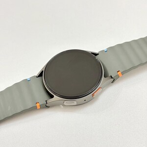 May include: A grey smartwatch with a round black screen and a grey strap. The strap has a textured design and is accented with blue and orange details. A modern tech accessory.