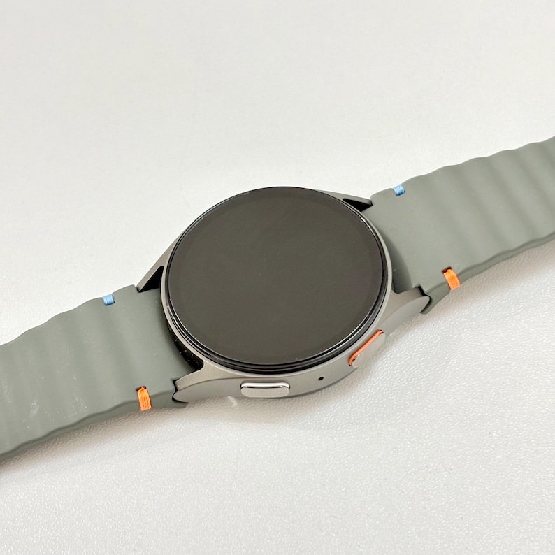 May include: A grey smartwatch with a round black screen and a grey strap. The strap has a textured design and is accented with blue and orange details. A modern tech accessory.