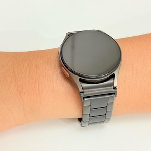 K&ouml;nnte beinhalten: Eine dunkelgraue Smartwatch mit rundem Zifferblatt und passendem Metallarmband wird am Handgelenk getragen. Die Uhr hat einen schwarzen Bildschirm und einen kleinen orangefarbenen Akzent. Das Armband besteht aus rechteckigen Gliedern.