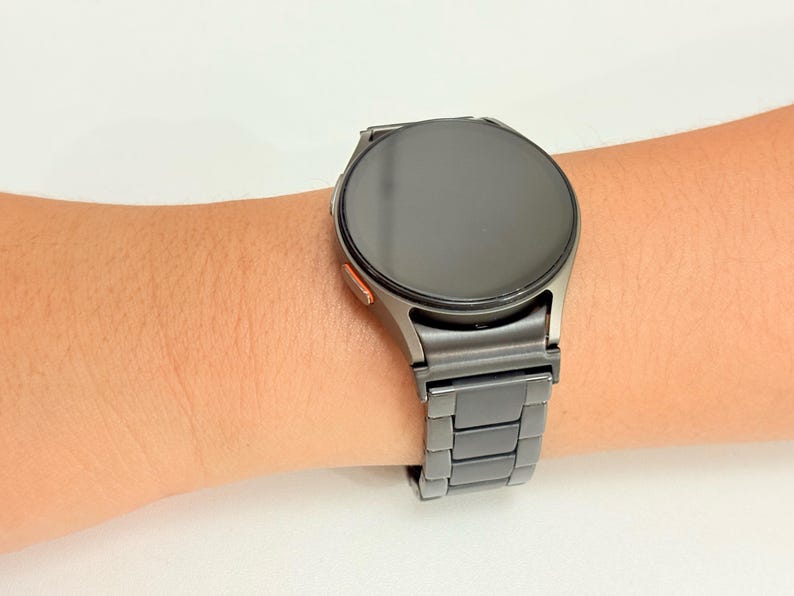 K&ouml;nnte beinhalten: Eine dunkelgraue Smartwatch mit rundem Zifferblatt und passendem Metallarmband wird am Handgelenk getragen. Die Uhr hat einen schwarzen Bildschirm und einen kleinen orangefarbenen Akzent. Das Armband besteht aus rechteckigen Gliedern.