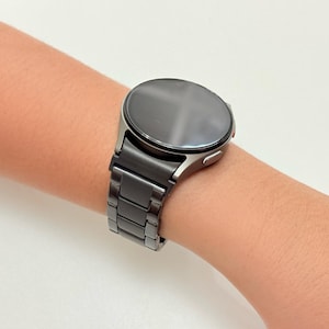 K&ouml;nnte beinhalten: Eine schwarze Smartwatch mit rundem Zifferblatt und passendem schwarzem Metallarmband wird am Handgelenk gezeigt. Die Uhr hat ein schlankes, modernes Design mit dunklem Bildschirm und poliertem Metallgeh&auml;use. Das Armband besteht aus rechteckigen Gliedern.