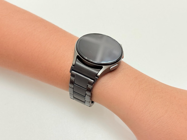 K&ouml;nnte beinhalten: Eine schwarze Smartwatch mit rundem Zifferblatt und passendem schwarzem Metallarmband wird am Handgelenk gezeigt. Die Uhr hat ein schlankes, modernes Design mit dunklem Bildschirm und poliertem Metallgeh&auml;use. Das Armband besteht aus rechteckigen Gliedern.
