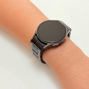 K&ouml;nnte beinhalten: Eine schwarze Smartwatch mit rundem Zifferblatt und passendem schwarzem Armband wird am Handgelenk getragen. Die Uhr hat ein schlankes, modernes Design mit dunklem Bildschirm und silberfarbener L&uuml;nette. Das Armband scheint aus Metall zu sein.