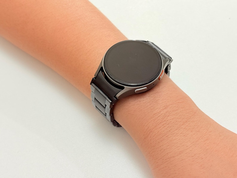 K&ouml;nnte beinhalten: Eine schwarze Smartwatch mit rundem Zifferblatt und passendem schwarzem Armband wird am Handgelenk getragen. Die Uhr hat ein schlankes, modernes Design mit dunklem Bildschirm und silberfarbener L&uuml;nette. Das Armband scheint aus Metall zu sein.