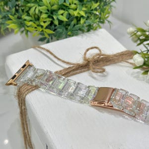 Könnte beinhalten: Ein klarer, schillernder Uhrenarmband mit roségoldener Schließe und Anschlüssen. Das Armband besteht aus rechteckigen, facettierten Gliedern, die das Licht einfangen. Das Uhrenarmband wird auf einer weißen Oberfläche mit einem natürlichen Bindfadenakzent präsentiert.