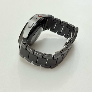 K&ouml;nnte beinhalten: Eine moderne Smartwatch mit einem schwarzen Metallarmband. Das Zifferblatt der Uhr ist silberfarben mit einem schwarzen Bildschirm. Auf der Seite ist ein kleiner roter Knopf zu sehen. Das Armband besteht aus rechteckigen Gliedern.