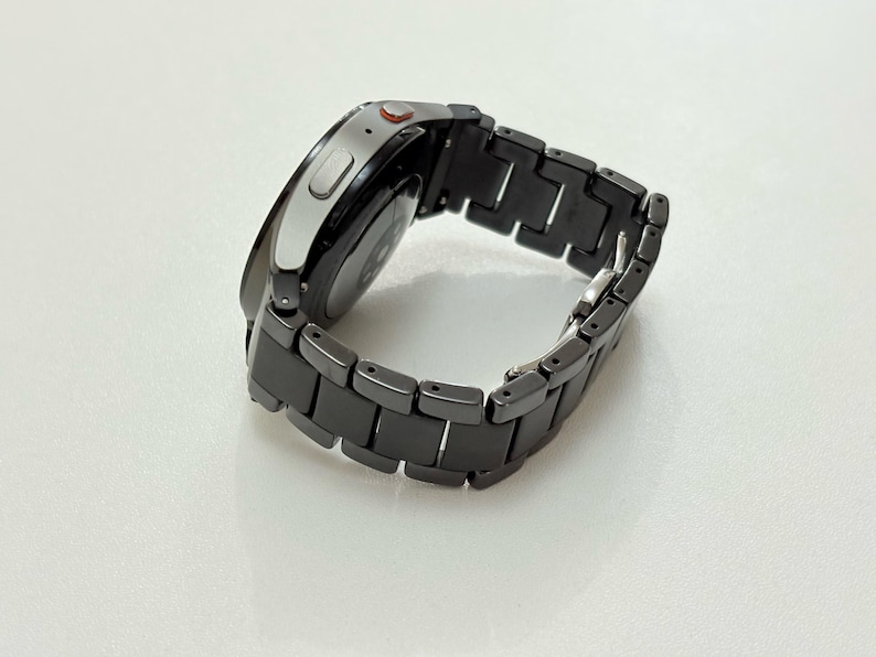K&ouml;nnte beinhalten: Eine moderne Smartwatch mit einem schwarzen Metallarmband. Das Zifferblatt der Uhr ist silberfarben mit einem schwarzen Bildschirm. Auf der Seite ist ein kleiner roter Knopf zu sehen. Das Armband besteht aus rechteckigen Gliedern.