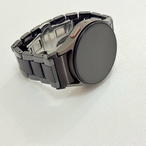 K&ouml;nnte beinhalten: Eine schwarze Smartwatch mit einem passenden schwarzen Metallarmband. Das Zifferblatt der Uhr ist rund und dunkel, mit einem kleinen orangefarbenen Akzent an der Seite. Das Armband besteht aus rechteckigen Gliedern.