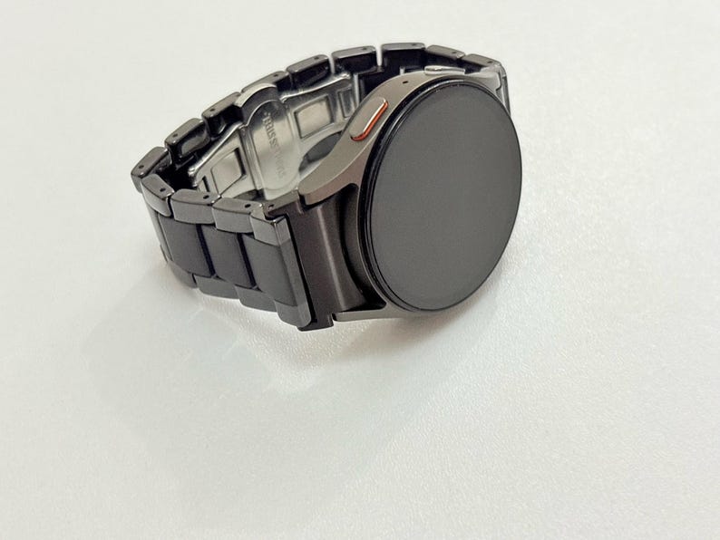 K&ouml;nnte beinhalten: Eine schwarze Smartwatch mit einem passenden schwarzen Metallarmband. Das Zifferblatt der Uhr ist rund und dunkel, mit einem kleinen orangefarbenen Akzent an der Seite. Das Armband besteht aus rechteckigen Gliedern.