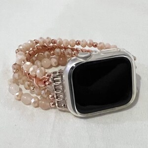 May include: An Apple Watch with a silver case and a beaded strap. The strap features multiple strands of light peach-coloured beads and rose gold-coloured metal accents. The watch face is black.