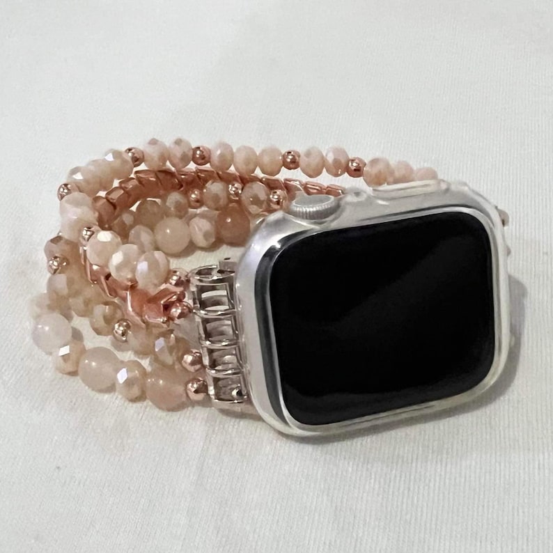 May include: An Apple Watch with a silver case and a beaded strap. The strap features multiple strands of light peach-coloured beads and rose gold-coloured metal accents. The watch face is black.