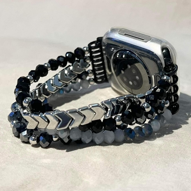 May include: A watch strap with a silver watch case. The strap features black, grey, and silver beads in various shapes and sizes. The strap is attached to a silver watch case.