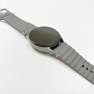 May include: A grey smartwatch with a round black screen and a grey ribbed silicone strap. The strap has orange and blue accent colours near the watch face. The watch is displayed on a white surface.
