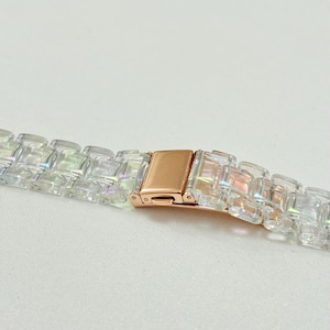 May include: Close-up of a clear, iridescent watch band with a rose gold clasp. The band is made of rectangular links that catch the light, creating a rainbow effect. The clasp is rectangular and has a secure closure.
