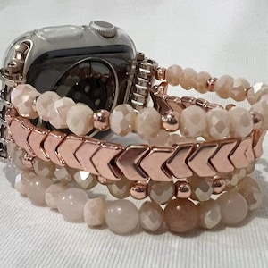 May include: An Apple Watch with a stack of beaded bracelets. The bracelets feature a mix of peach-coloured faceted beads and rose gold chevron-shaped beads. The watch has a silver case and is attached to the bracelets.