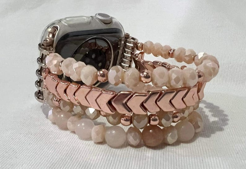 May include: An Apple Watch with a stack of beaded bracelets. The bracelets feature a mix of peach-coloured faceted beads and rose gold chevron-shaped beads. The watch has a silver case and is attached to the bracelets.