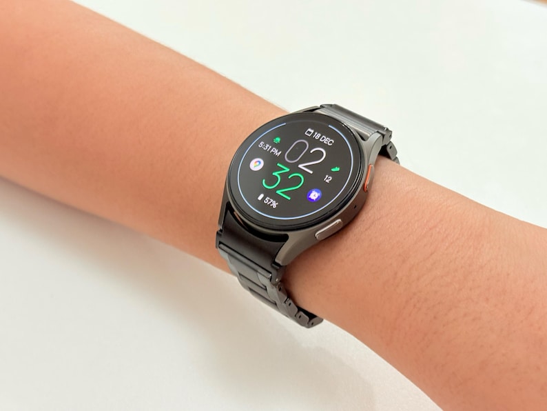 K&ouml;nnte beinhalten: Eine schwarze Smartwatch mit Metallarmband wird am Handgelenk gezeigt. Das Zifferblatt zeigt die Uhrzeit 02:32 an, zusammen mit anderen Symbolen und dem Datum 18. Dezember. Der Akkuladestand betr&auml;gt 57%.