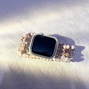 May include: A silver smartwatch with a black screen and a beaded strap. The strap features multiple strands of small, light pink and white beads, with silver and rose gold accents. The watch is on a white surface.