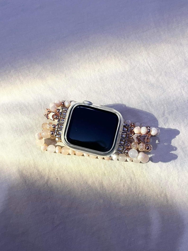 May include: A silver smartwatch with a black screen and a beaded strap. The strap features multiple strands of small, light pink and white beads, with silver and rose gold accents. The watch is on a white surface.