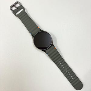 May include: A grey smartwatch with a dark screen and a matching grey strap. The strap has a ribbed texture and a metal buckle. Small orange and blue accents are visible on the strap near the watch face.