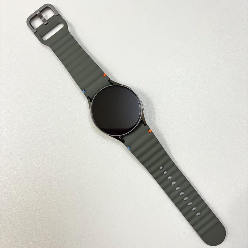 May include: A grey smartwatch with a dark screen and a matching grey strap. The strap has a ribbed texture and a metal buckle. Small orange and blue accents are visible on the strap near the watch face.