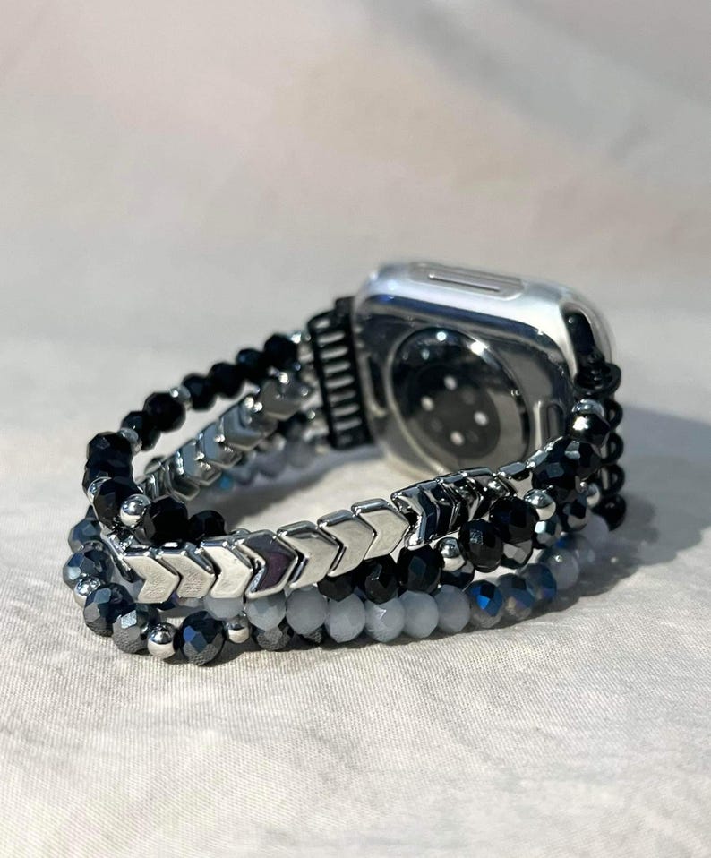 May include: Silver Apple Watch with a beaded strap. The strap features black, grey, and silver beads in a layered design. The strap has a mix of round and arrow-shaped beads.